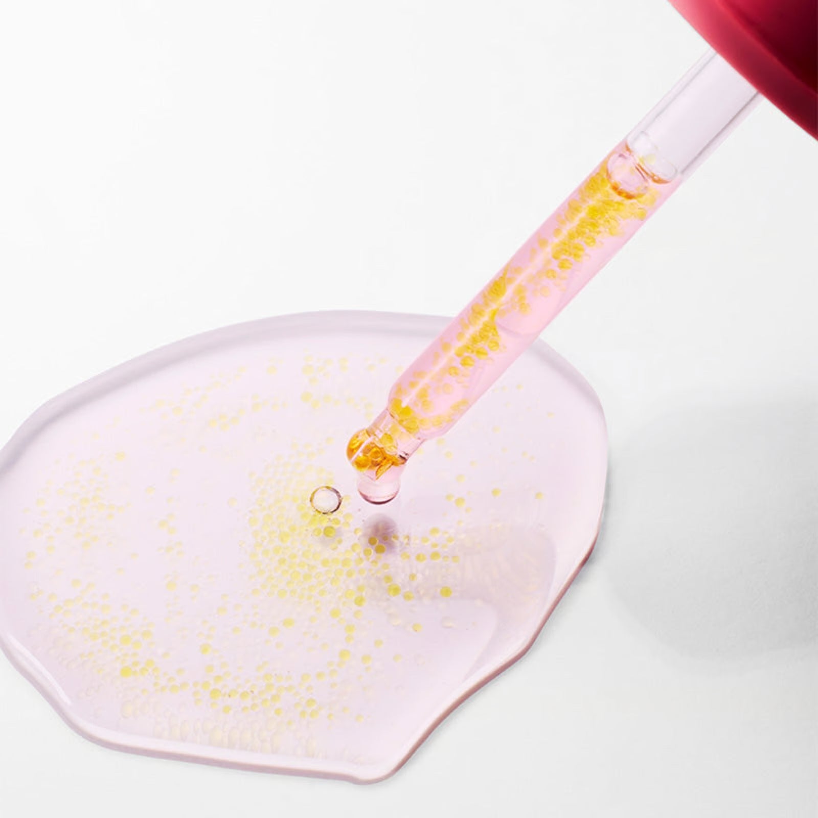 Pink serum being dispensed from a glass dropper onto a surface, highlighting the retinal beads.