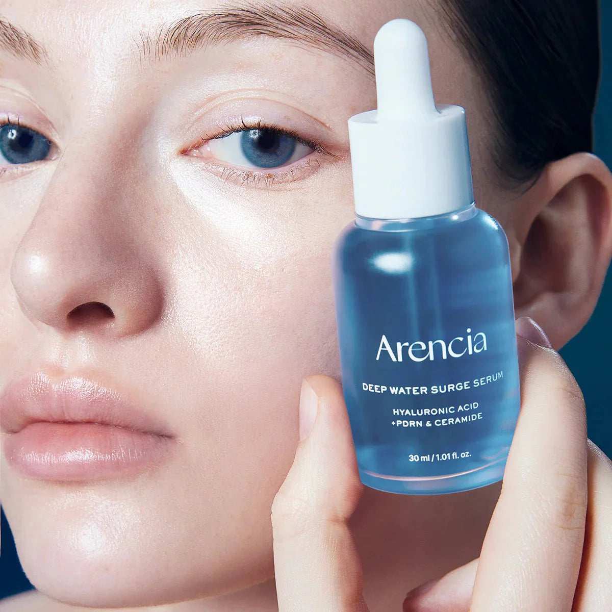 Close-up of a model with glowing, dewy skin holding the Arencia Deep Water Surge Serum bottle near her face.