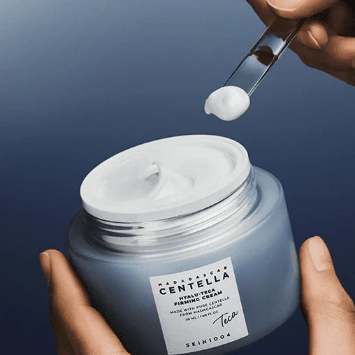 A hand using a clear spatula to scoop Skin1004 Hyalu-Teca Firming Cream from the blue frosted jar.