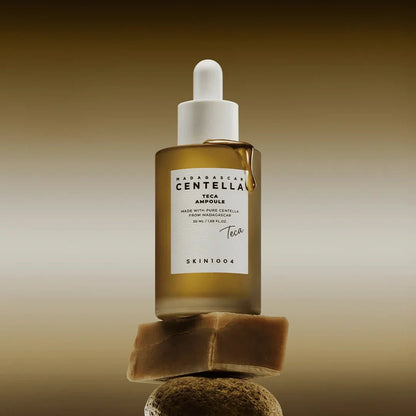 Skin1004 Centella Teca Ampoule bottle balanced on a natural stone with a golden liquid drip.
